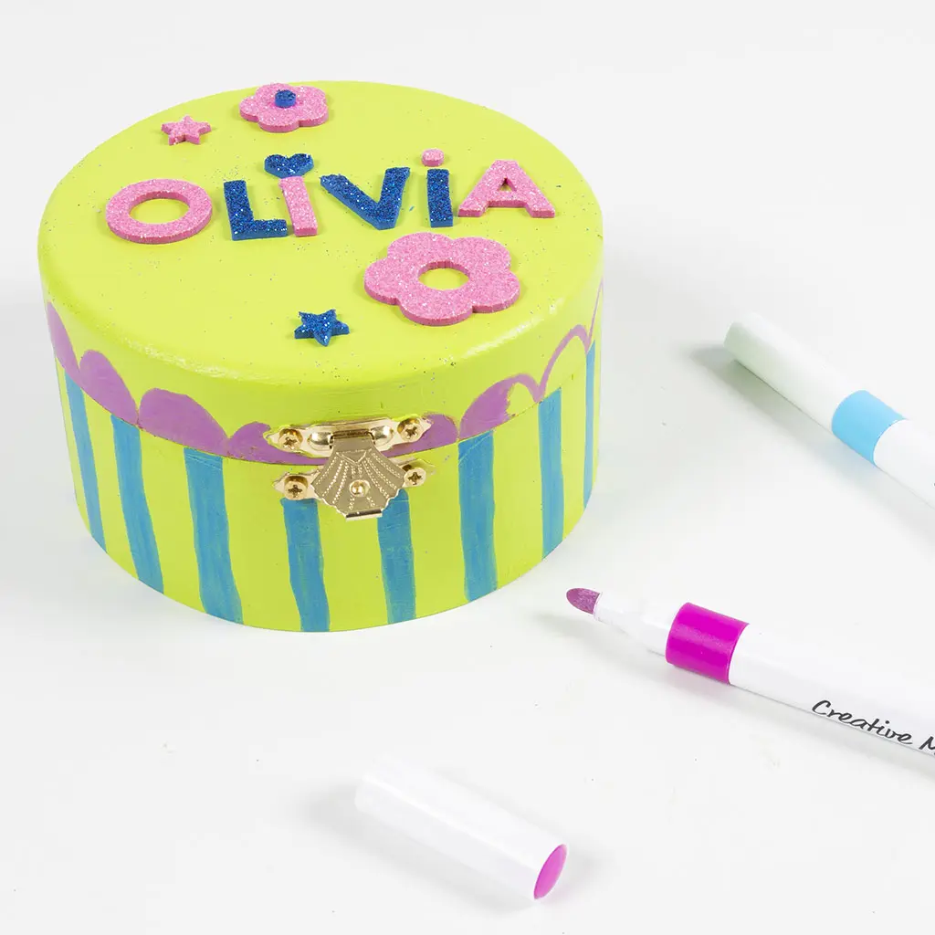 Close-up van het afgewerkte gele kistje met de naam Olivia in glitter foam letters, bloemen en sterren op het deksel. De zijkant is beschilderd met een paars-blauw patroon van bogen en strepen. Ernaast liggen stiften in wit, lichtblauw en roze/paars.