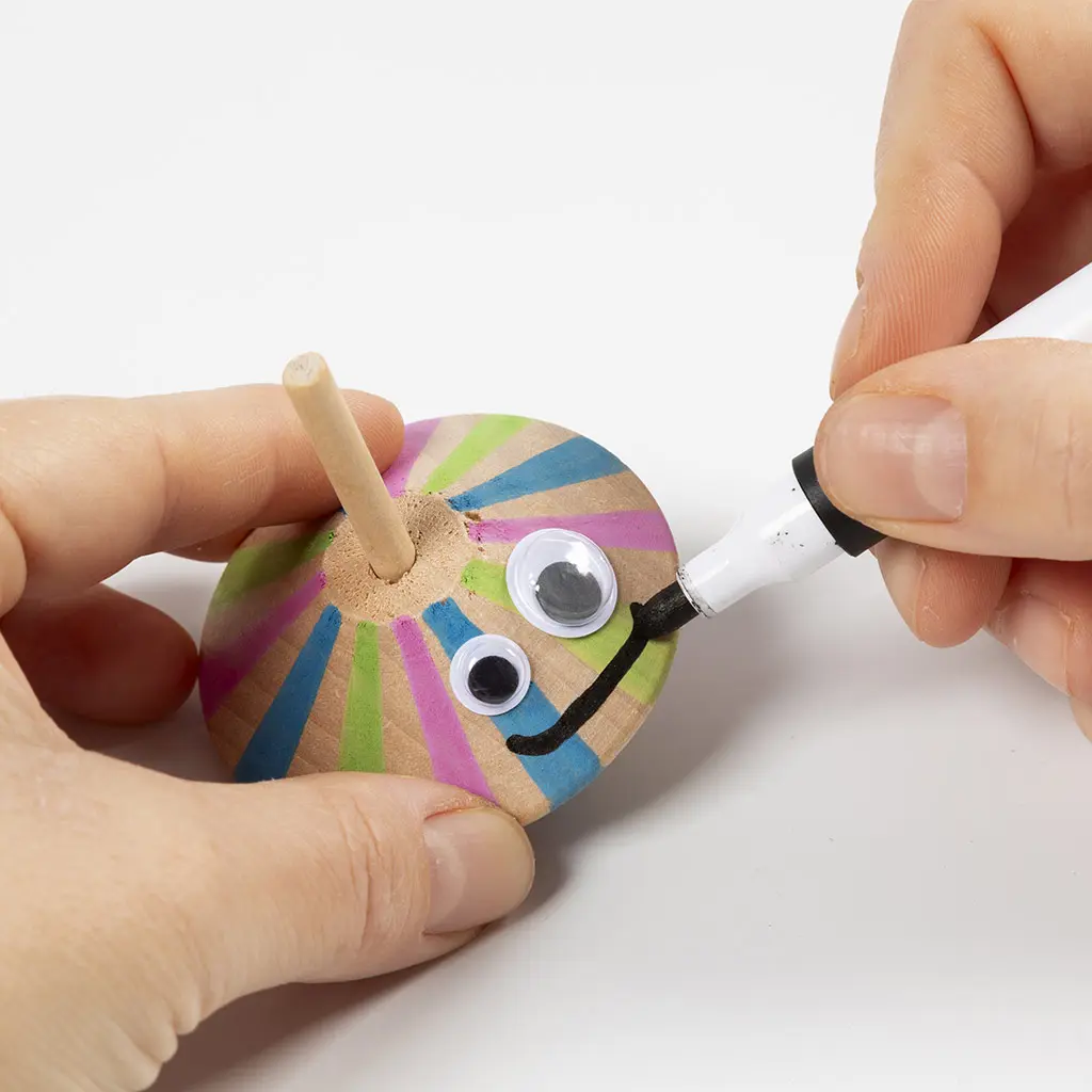 Met een zwarte marker wordt er een glimlach getekend onder de wiebeloogjes op de houten tol.