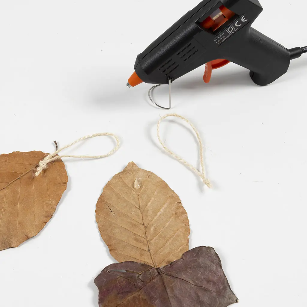 Een lijmpistool boven een gedroogd herfstblad waarop een drup warmelijm is aangebracht om een touwlusje vast te zetten.