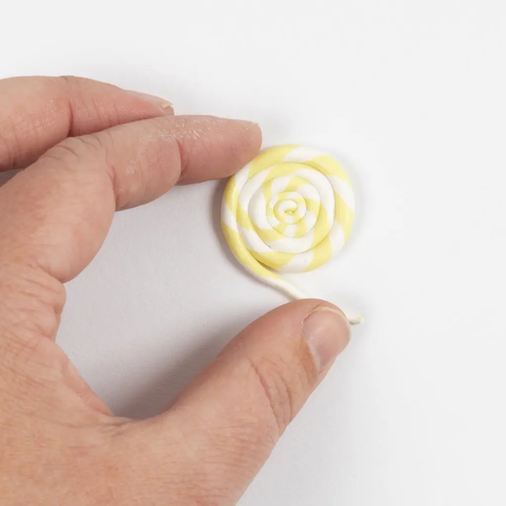 Twee handen rollen de geel-wit gestreepte kleistring op tot een ronde schijf voor een spiraalsnoepje of lolly.