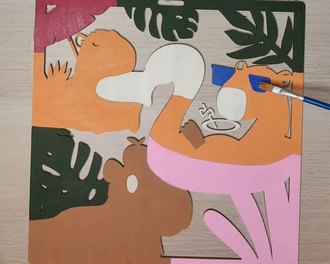 Houten laag gedeeltelijk ingekleurd in oranje, roze, wit en donkergroen met een penseel, met capybara- en bladervormen zichtbaar.