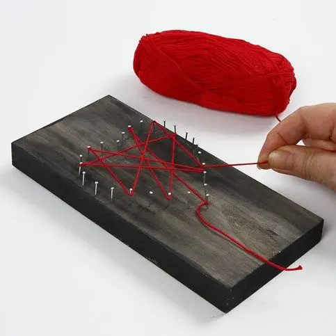 Een hand begint een rode draad kriskras tussen de spijkers op de houten plank te spannen (string art). Rechtsboven ligt een bol rode wol.