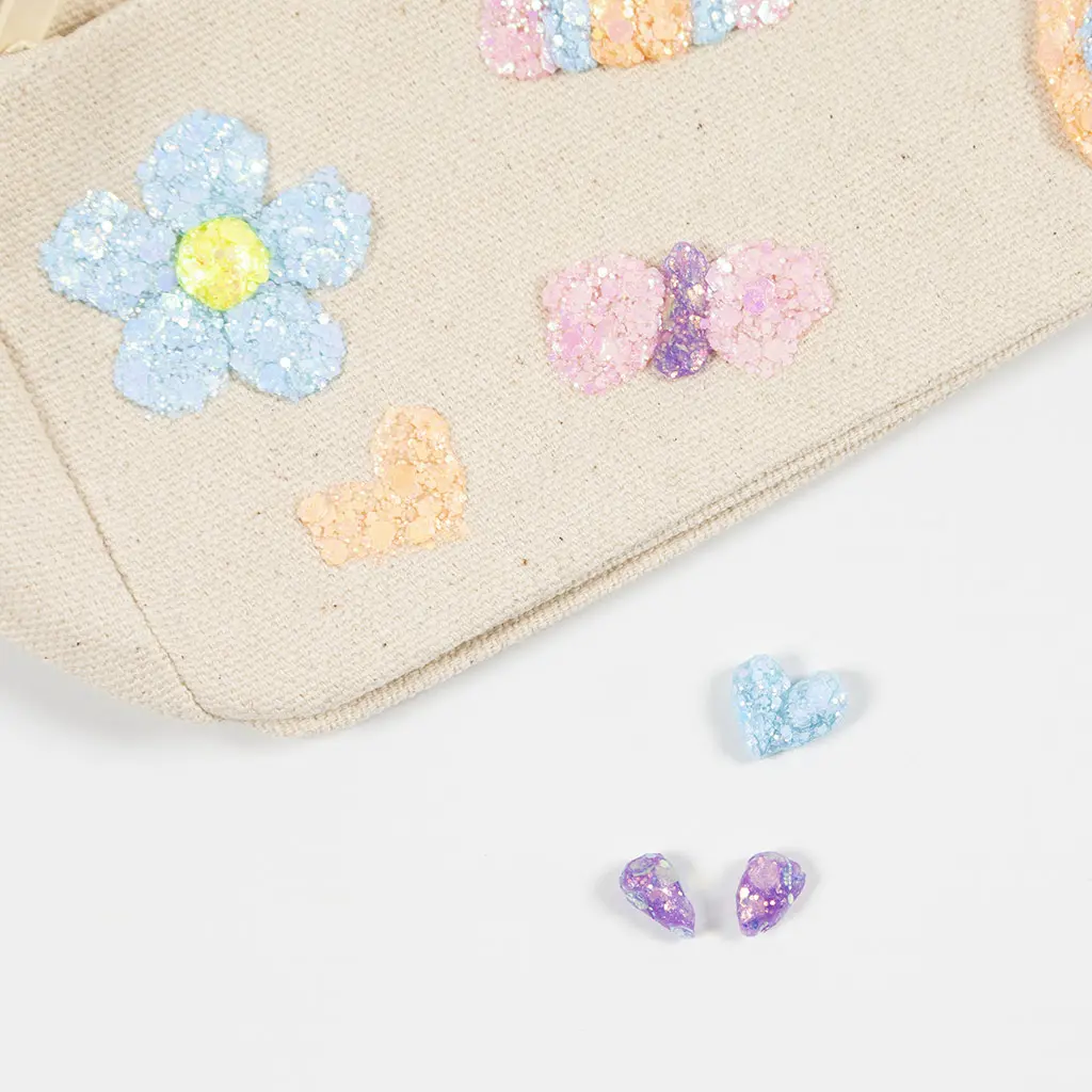 Detailopname van een canvas etui met een blauw glitter bloemetje, een oranje hartje en een roze-paarse strik, naast twee losse blauwe en paarse glitter hartjes.