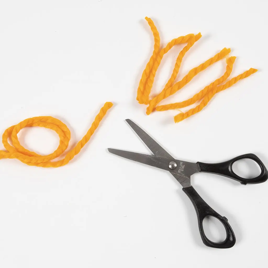 Een zwarte knutselschaar ligt op een witte achtergrond naast feloranje garen, waarvan er een aantal draden in korte stukjes zijn geknipt.