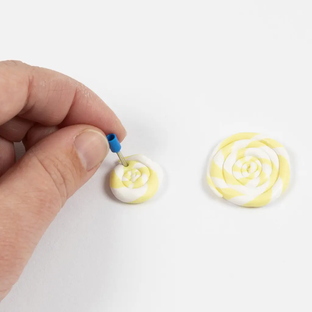 Een hand gebruikt een klein blauw gereedschapje om een gat te maken in het midden van een klein geel-wit opgerold kleisnoepje, met een grotere lolly erachter.