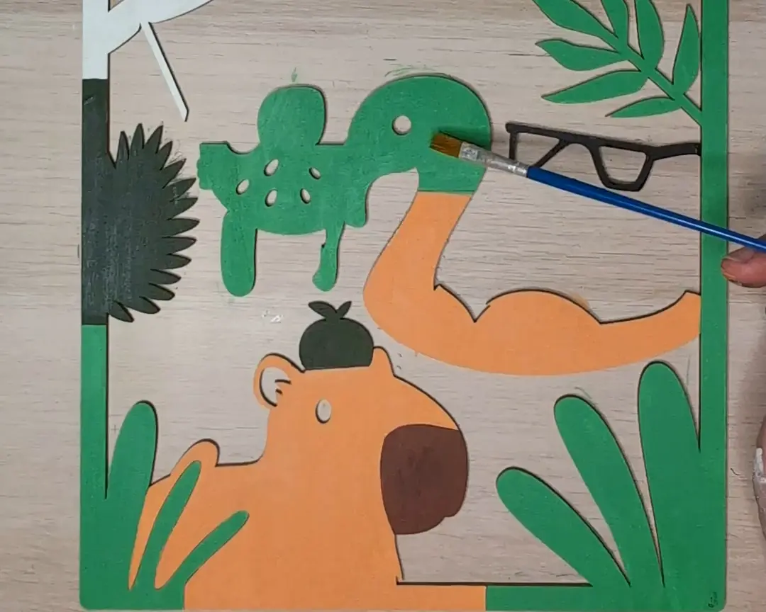 Houten laag met een capybara met fruit op zijn hoofd en groene plantvormen, ingekleurd in oranje, groen en bruin met een penseel.