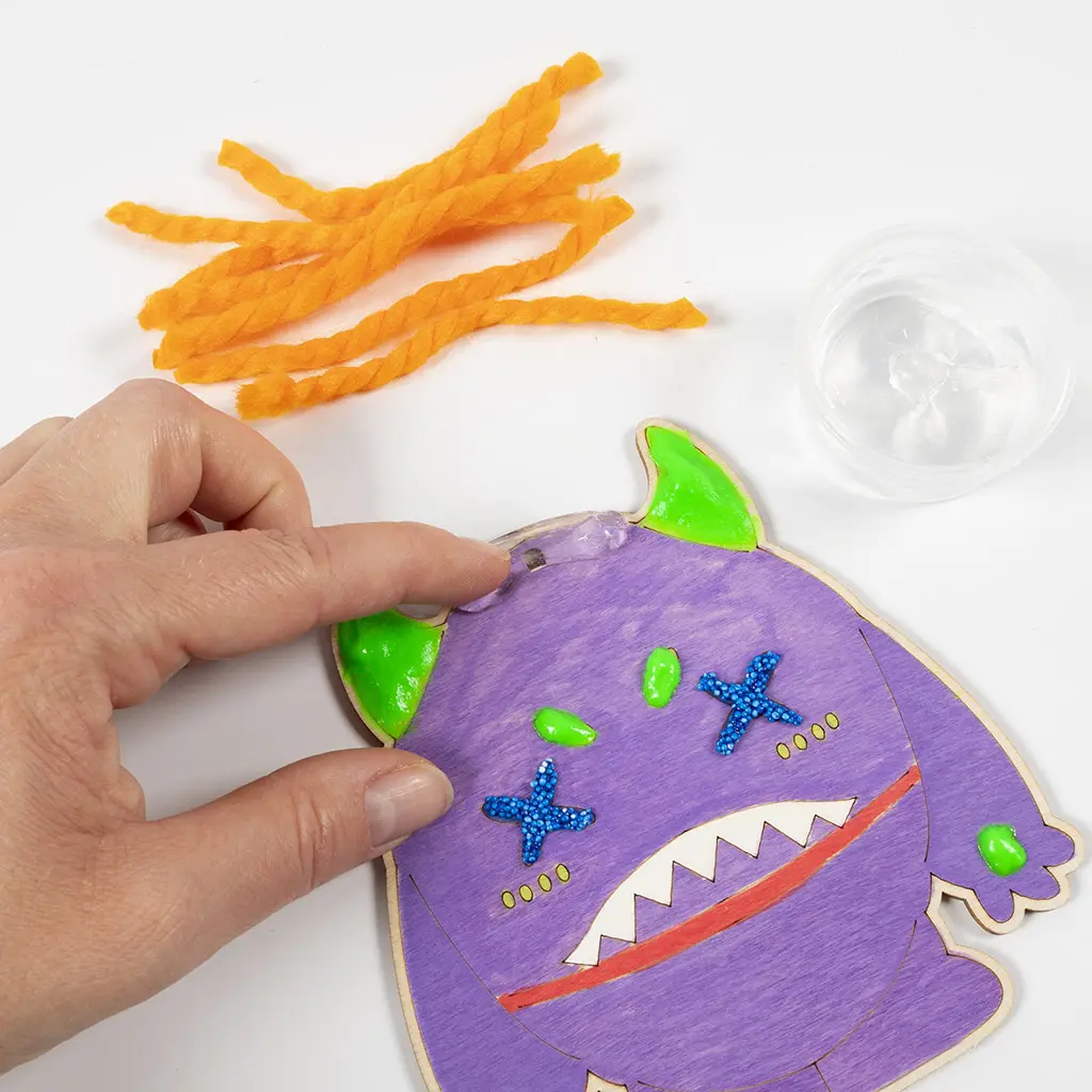 Een hand brengt transparante hobbylijm aan op de bovenrand tussen de hoorns van het paarse houten monster. Op de achtergrond liggen de stukjes oranje wol en een klein plastic bakje met lijm.