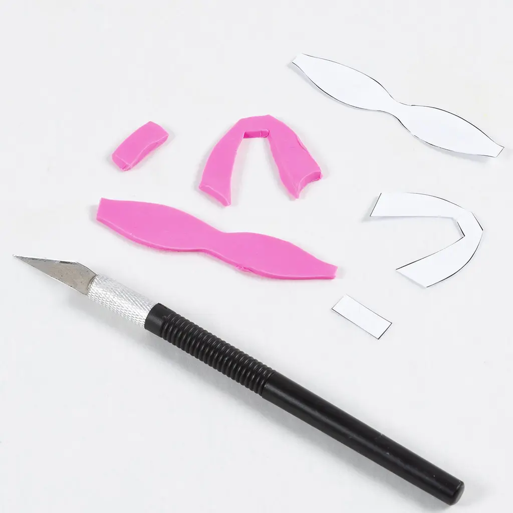 Uitgeknipte papieren sjablonen voor een strik liggen naast de bijbehorende uitgesneden roze kleionderdelen op een witte ondergrond, met een zilverkleurig hobbymes op de voorgrond.