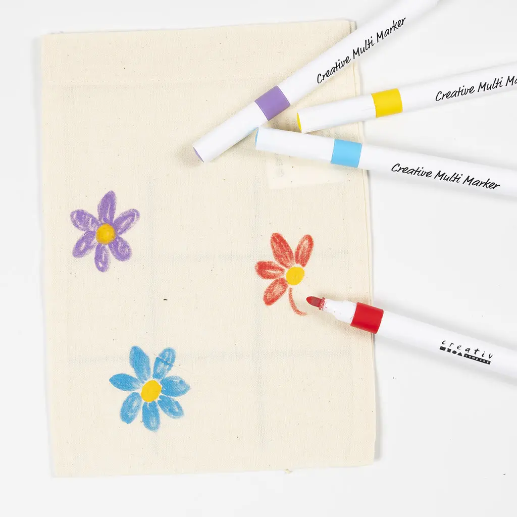 De achterkant van het stoffen zakje wordt versierd met kleurrijke bloemen. Er zijn al een paarse en een blauwe bloem getekend en er wordt met een rode marker een derde bloem ingekleurd. Er liggen nog drie andere markers bovenaan de tas.