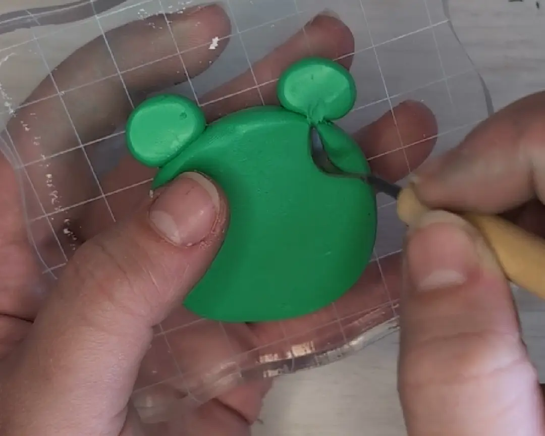 Een hand gebruikt een houten boetseerspatel met een platte, metalen kop om de basis van de twee groene kikkerogen glad te strijken en te laten overvloeien in het kikkellijfje.