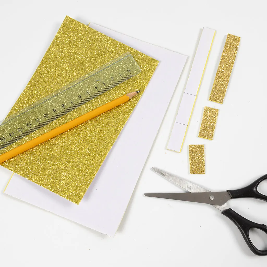 Een vel gouden glitterpapier met een liniaal en potlood erop, naast afgesneden gouden glitterstroken en een schaar.