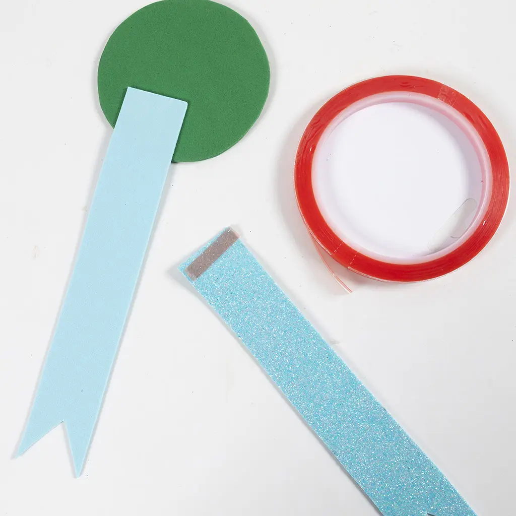 Een groene foam cirkel, een lichtblauwe foam strook met gespleten puntje, een lichtblauwe glitter strook en een rol dubbelzijdig tape op een witte ondergrond.