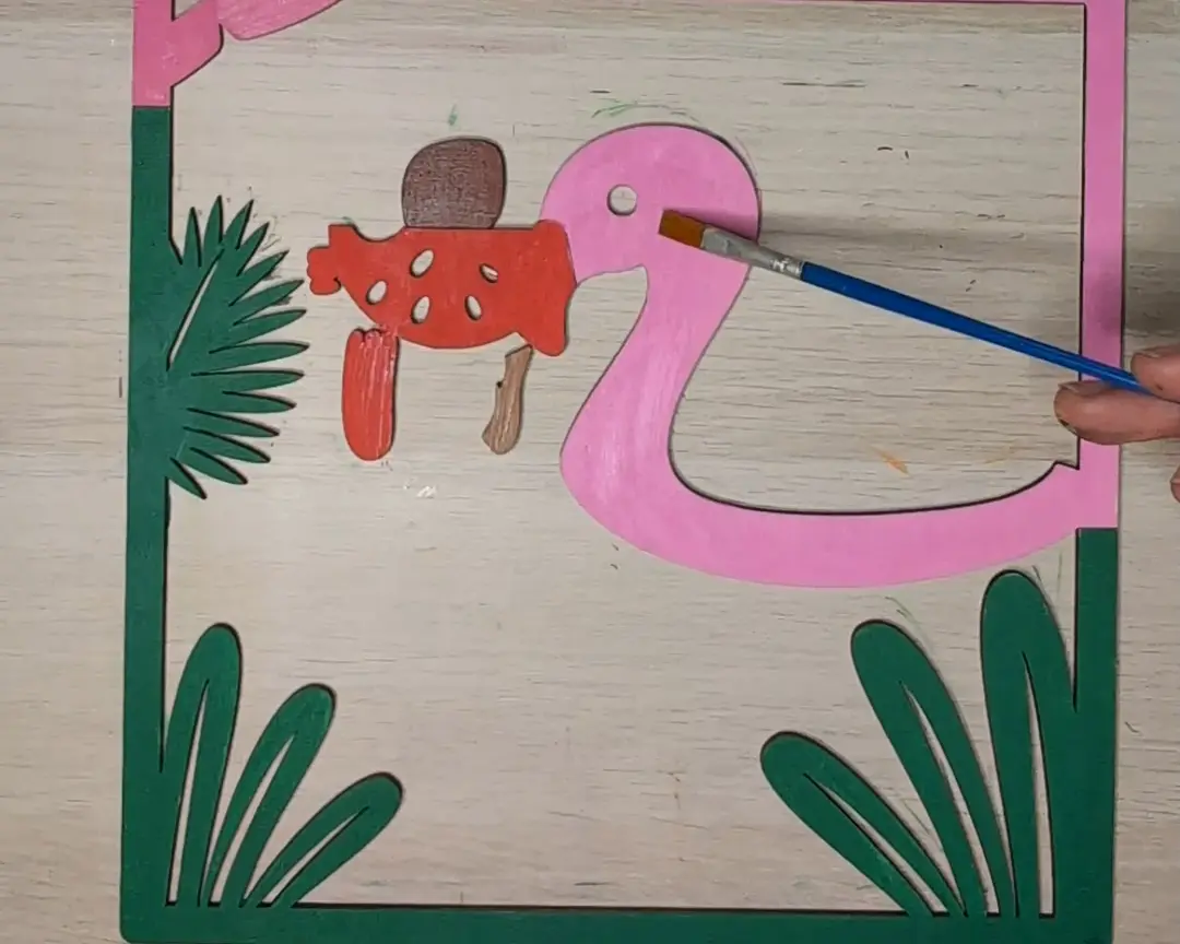 Houten laag met een roze flamingo en een rode decoratieve figuur, ingekleurd met een penseel tegen een achtergrond van groene bladvormen.