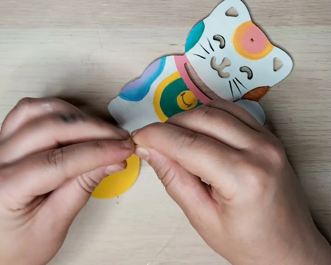 Handen die een doorzichtig draadje door het gaatje van de beschilderde maneki-neko figuur rijgen om de zonnevanger samen te stellen.