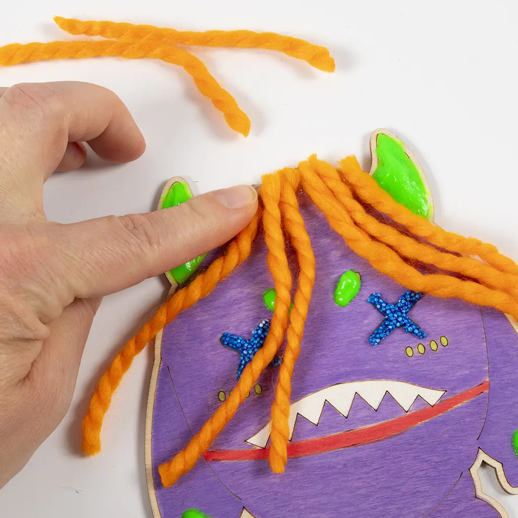 Een hand drukt de kortgeknipte plukjes oranje wol in de lijm op de bovenkant van het paarse monster om er een kapsel van te maken.