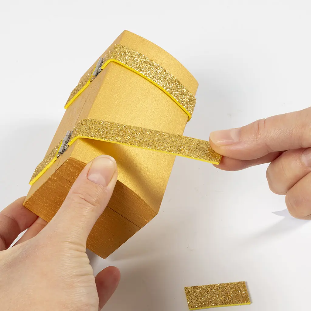 Twee handen plakken een zelfklevende gouden glitterstrook langs de rand van een goud geschilderd houten kistje met koepeldeksel.