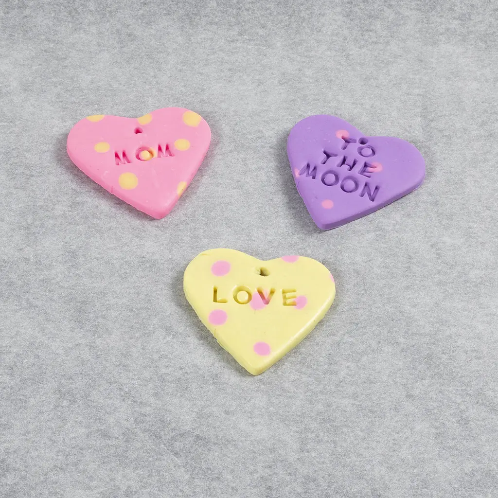 Drie gebakken kleihartjes met stippen en teksten zoals MOM, TO THE MOON en LOVE op bakpapier.