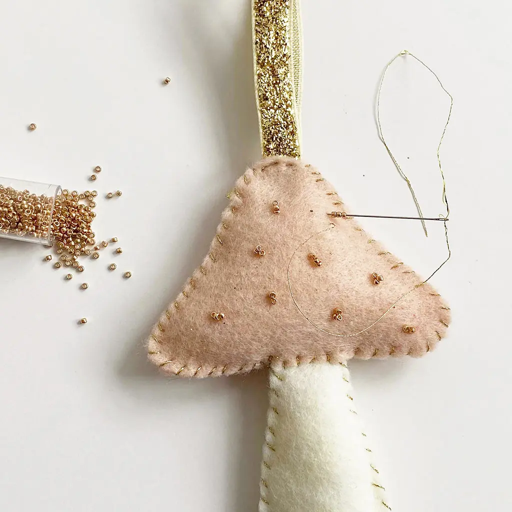 Een handgewerkte vilten paddenstoel hanger waarbij kleine gouden kraaltjes op het roze hoedje worden genaaid met naald en garen.