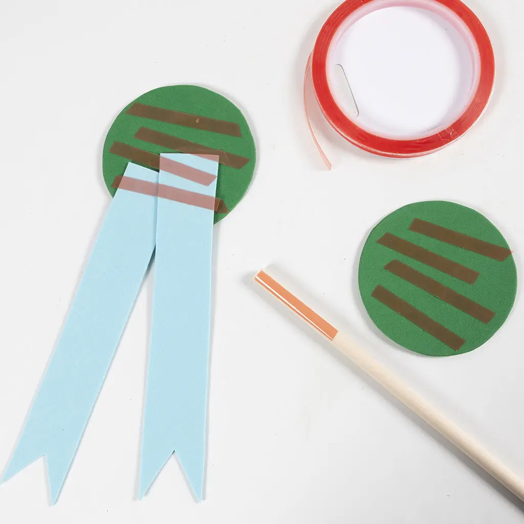 Twee groene foam cirkels met dubbelzijdig tape, lichtblauwe foam linten vastgeplakt aan één cirkel, een houten stokje en een rol dubbelzijdig tape op een witte ondergrond.