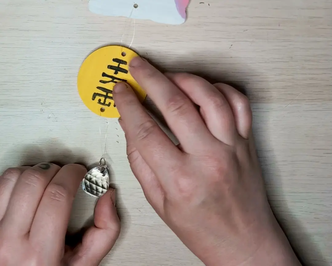 Handen die een transparant draadje bevestigen aan de gele ronde schijf met Japanse tekens, met daaraan een kleine glazen kristallen hanger.