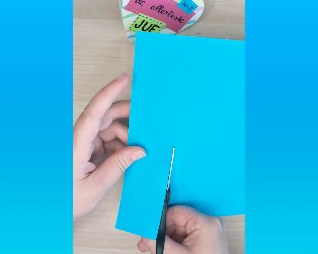 Een hand knipt een lichtblauwe papieren strook op maat met een schaar, met het afgewerkte hartdeksel zichtbaar op de achtergrond.