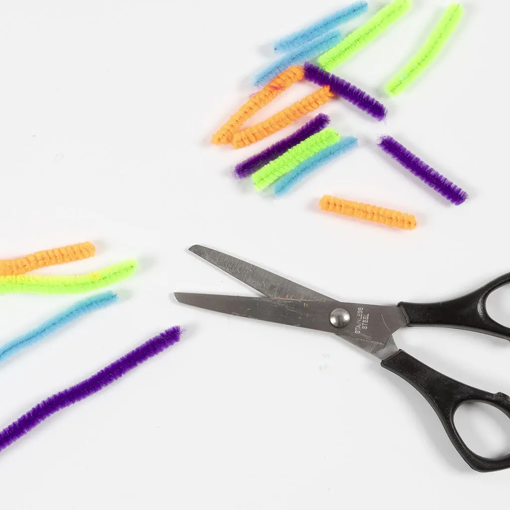 Een zwarte schaar ligt op een witte ondergrond tussen verschillende kortgeknipte stukjes kleurrijke chenilledraad (pijpenragers) in de kleuren paars, groen, blauw en oranje.