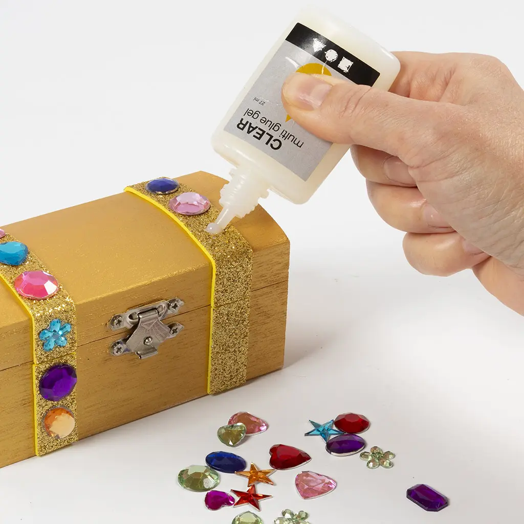 Een hand knijpt Clear multi glue gel uit een flesje op een goud geschilderd kistje met glitterstroken en gekleurde decoratiestenen, met losse stenen in verschillende vormen en kleuren naast het kistje.