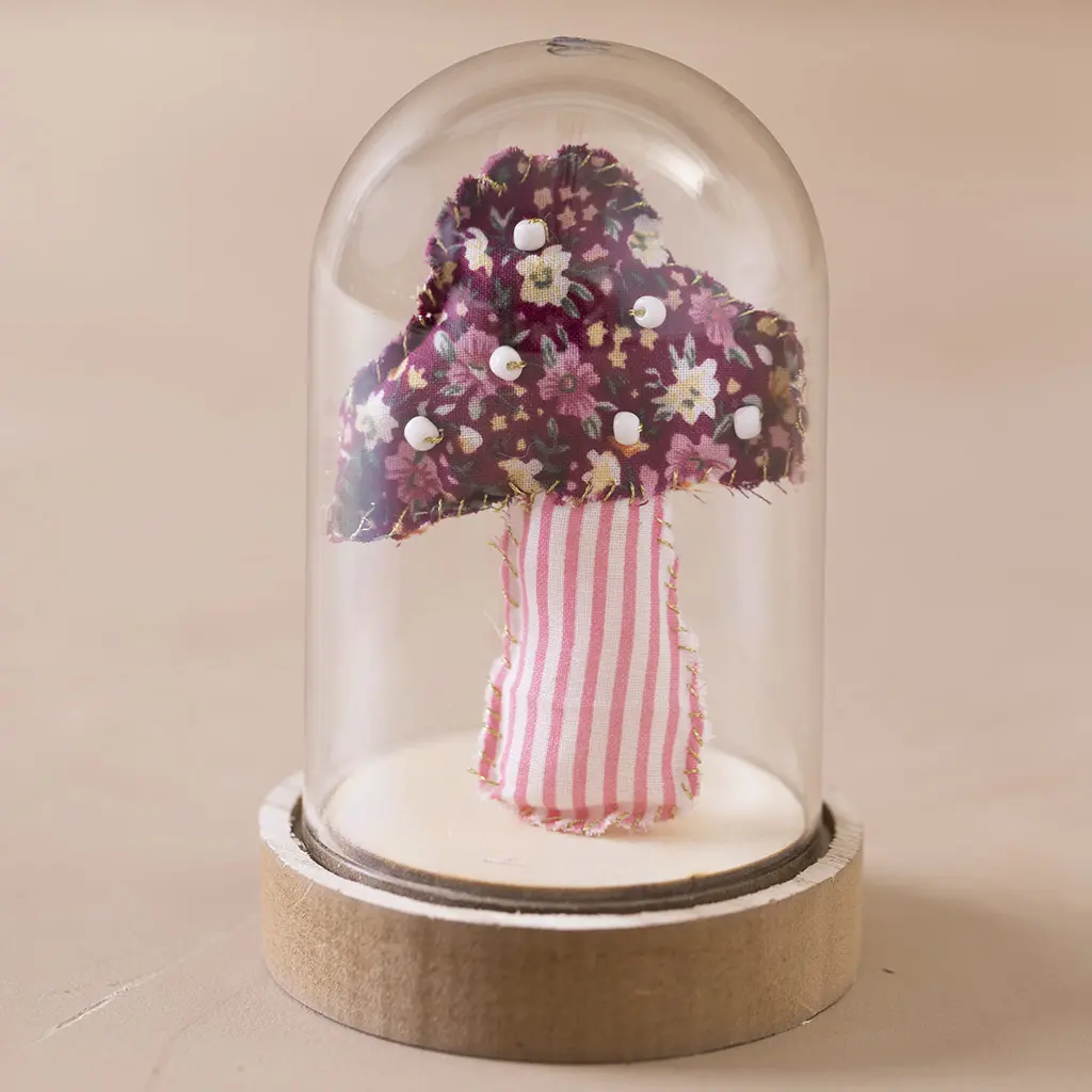 Een stoffen paddenstoeltje met een gebloemd hoedje en gestreept steelje, gedecoreerd met witte kraaltjes, gepresenteerd onder een glazen stoofje op een houten voet.