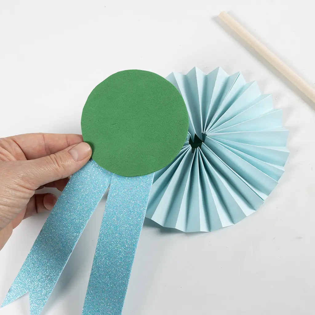 Een hand houdt een groene foam cirkel met lichtblauwe glitter linten, voor een lichtblauwe gevouwen papieren waaier rozet en een houten stokje op een witte ondergrond.