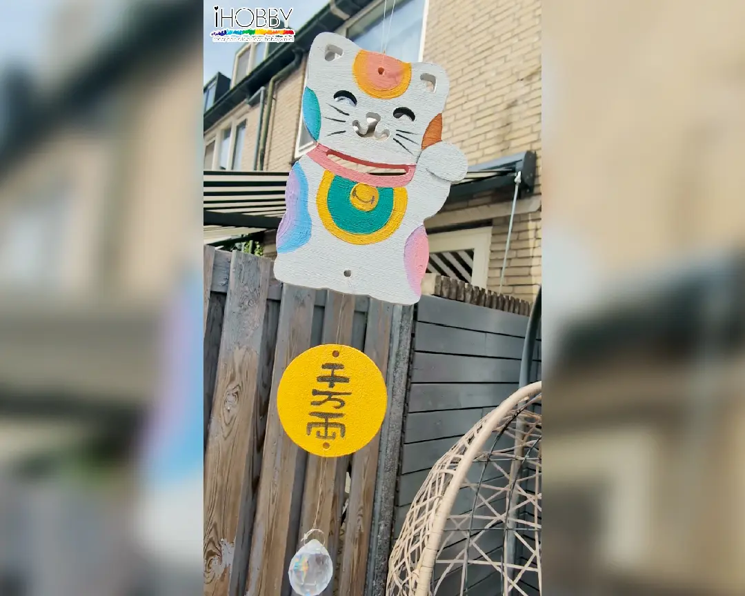 Afgewerkte zelfgemaakte zonnevanger hangend buiten in een tuin, bestaande van een beschilderde maneki-neko, een gele ronde schijf met Japanse tekens en een kristallen hanger.