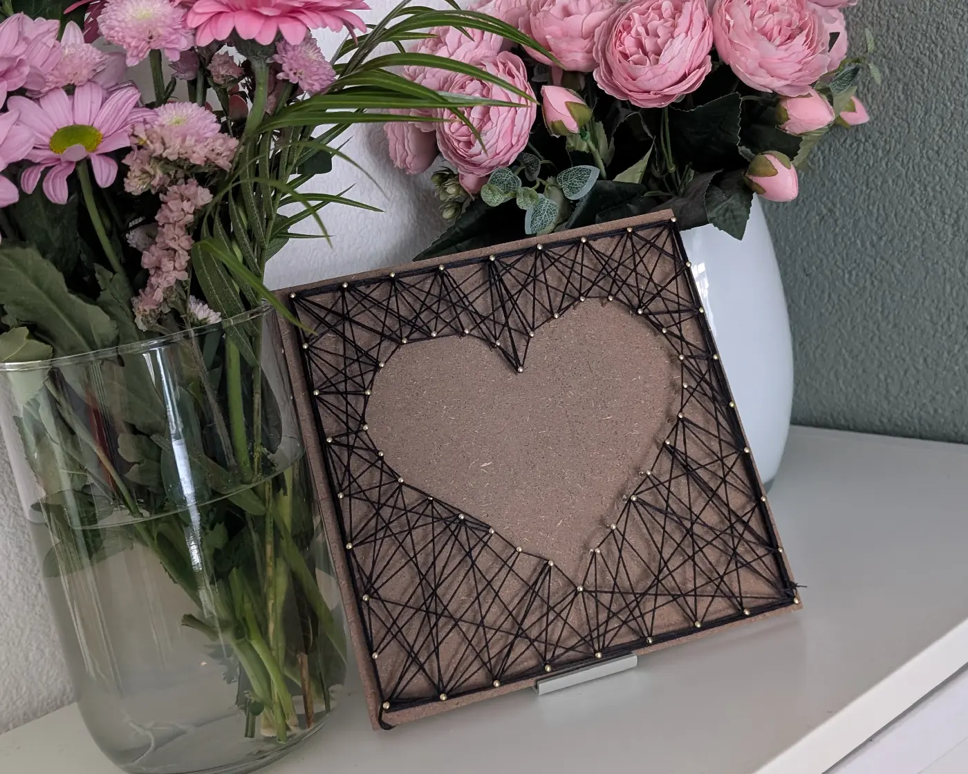 Afgewerkt string art hart van zwart garen op een houten plaat, tentoongesteld naast vazen met roze bloemen