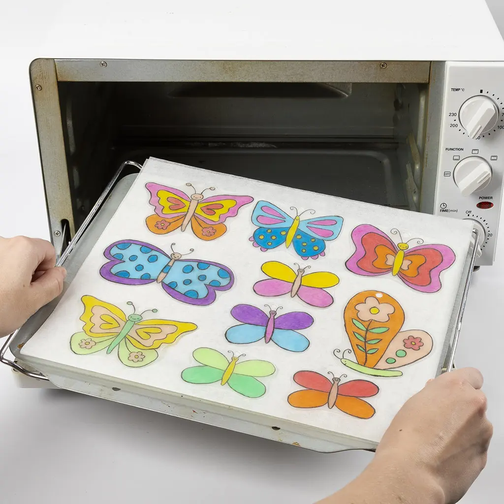 Twee handen schuiven een bakplaat met de krimpfolievlinders op een vel bakpapier in een kleine, witte elektrische oven.