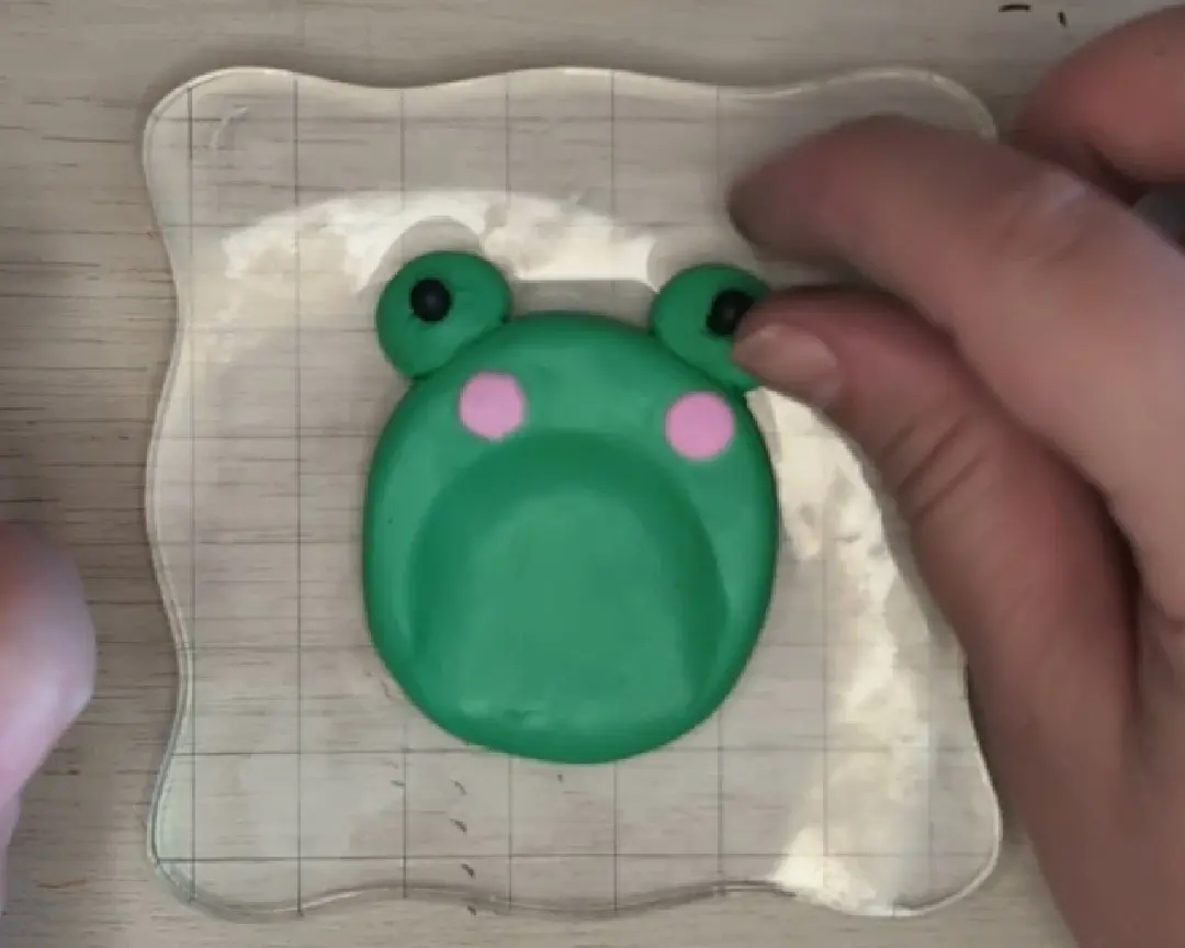 Een hand gebruikt een fijne metalen spatel om de kleine zwarte kleipupil in de oogkas van de kikker te drukken. Het andere oogje is al voorzien van een zwarte pupil.