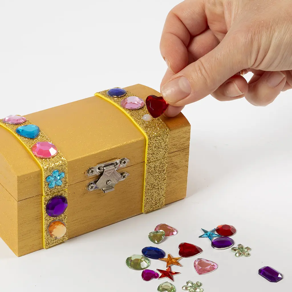 Een hand plaatst een rood hartje decoratiesteen op een goud geschilderd kistje met glitterstroken en kleurrijke decoratiestenen, met losse stenen naast het kistje.