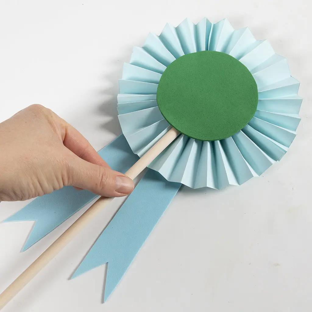 Een hand houdt een houten stokje vast waaraan een rozet is bevestigd met een lichtblauwe gevouwen papieren waaier, een groene foam cirkel in het midden en lichtblauwe foam linten onderaan.