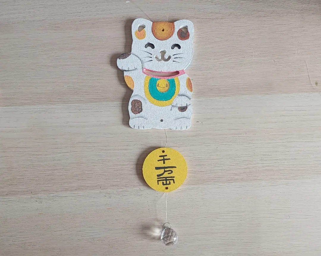 Afgewerkte zelfgemaakte zonnevanger op een houten ondergrond, bestaande uit een beschilderde MDF maneki-neko met roze halsband en gele munt, verbonden via een transparant draadje aan een gele ronde schijf met Japanse tekens en een kristallen hanger onderaan.