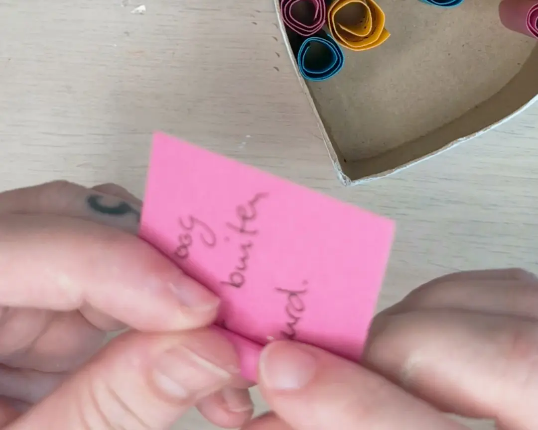 Twee handen rollen een beschreven roze kaartje op, met op de achtergrond een hartvormige doos gevuld met opgerolde kleurrijke kaartjes.
