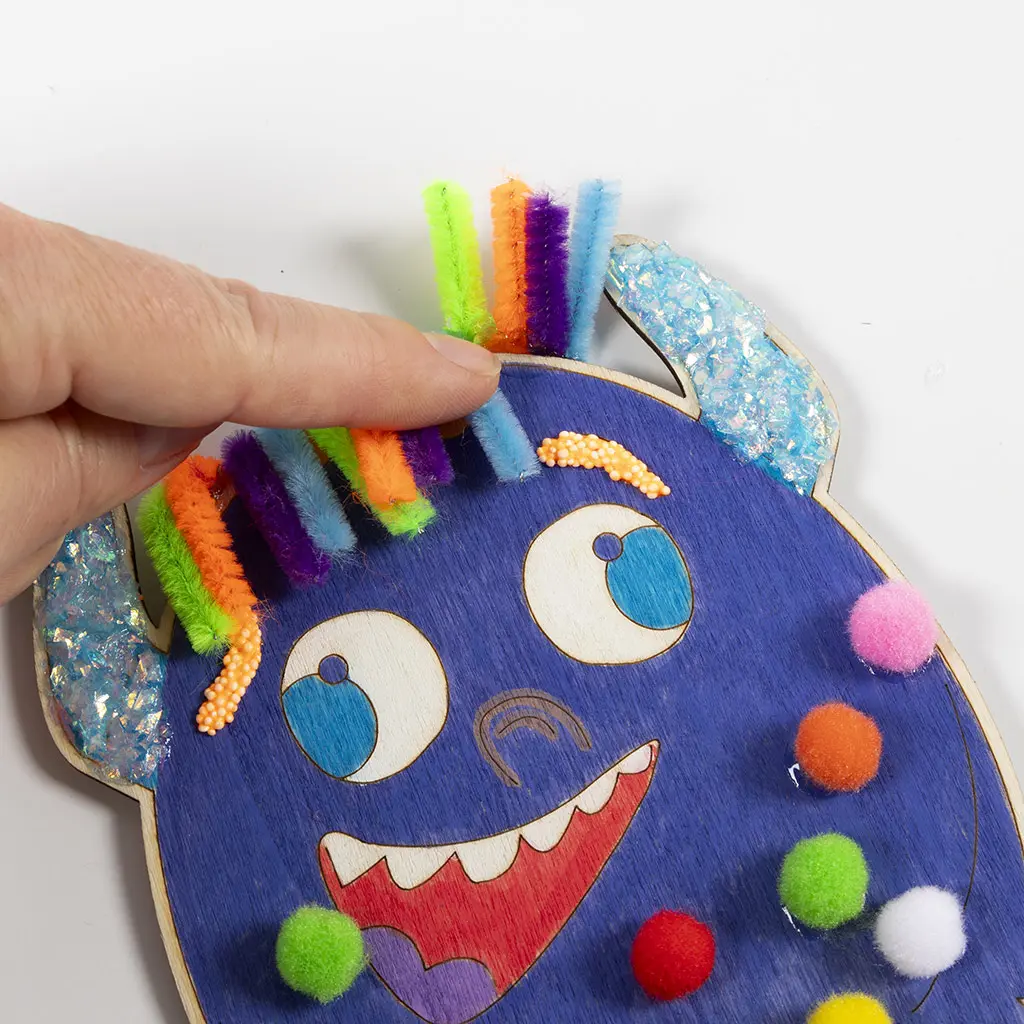 Een hand schikt de kleurrijke, rechtopstaande stukjes chenilledraad op het hoofd van het blauwe monster voor een vrolijk spikykapsel. Het monster is nu compleet met glitterhoorns, klei-wenkbrauwen en pompons.