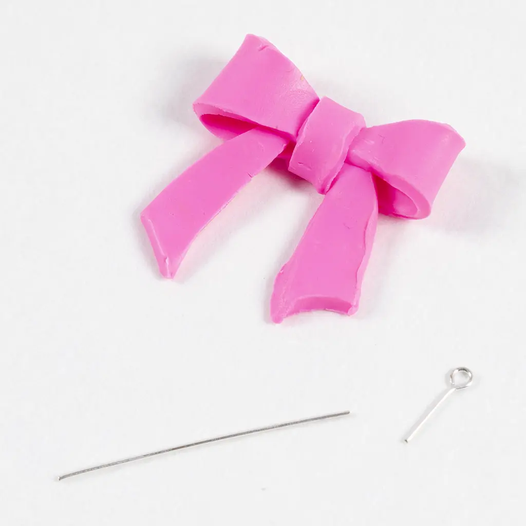 De voltooide roze kleistrik ligt schuin op een witte achtergrond. Eronder liggen een rechte metalen pin en een metalen oogpen die klaarstaan om in de strik te worden geschoven.