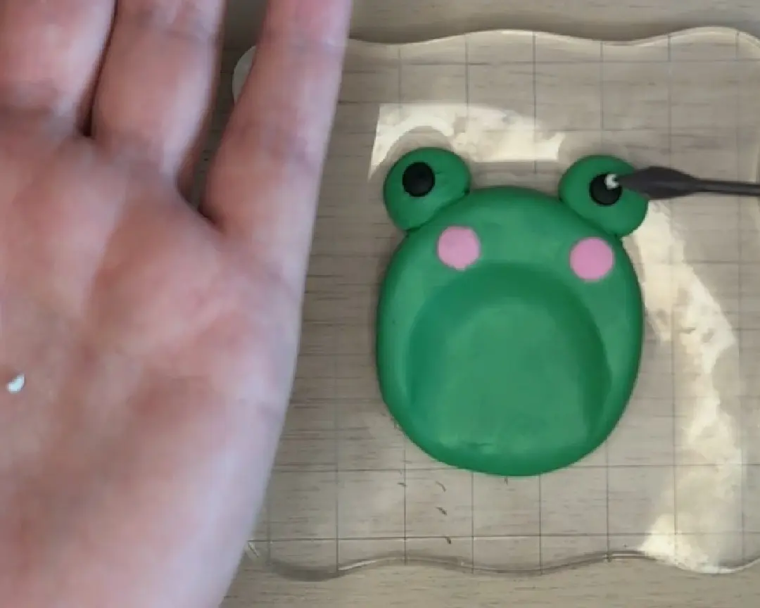 Een kikkergezichtje van groene klei ligt op het ruitjesblok. Met een fijne tool wordt een minuscuul wit bolletje klei als lichtreflectie op de zwarte pupil van de kikker geplaatst.