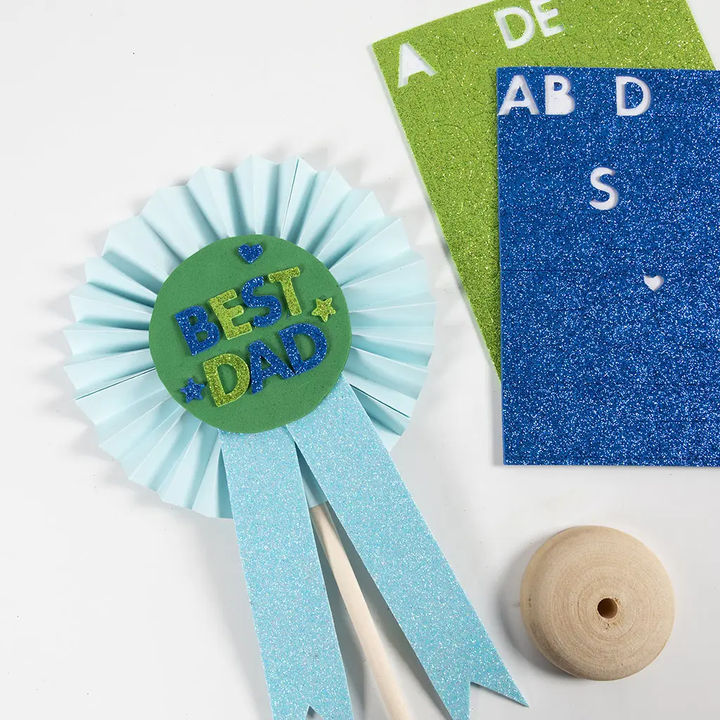 Een afgewerkte rozet op een houten stokje met lichtblauwe waaier, glitter linten en een groene foam cirkel met de tekst 