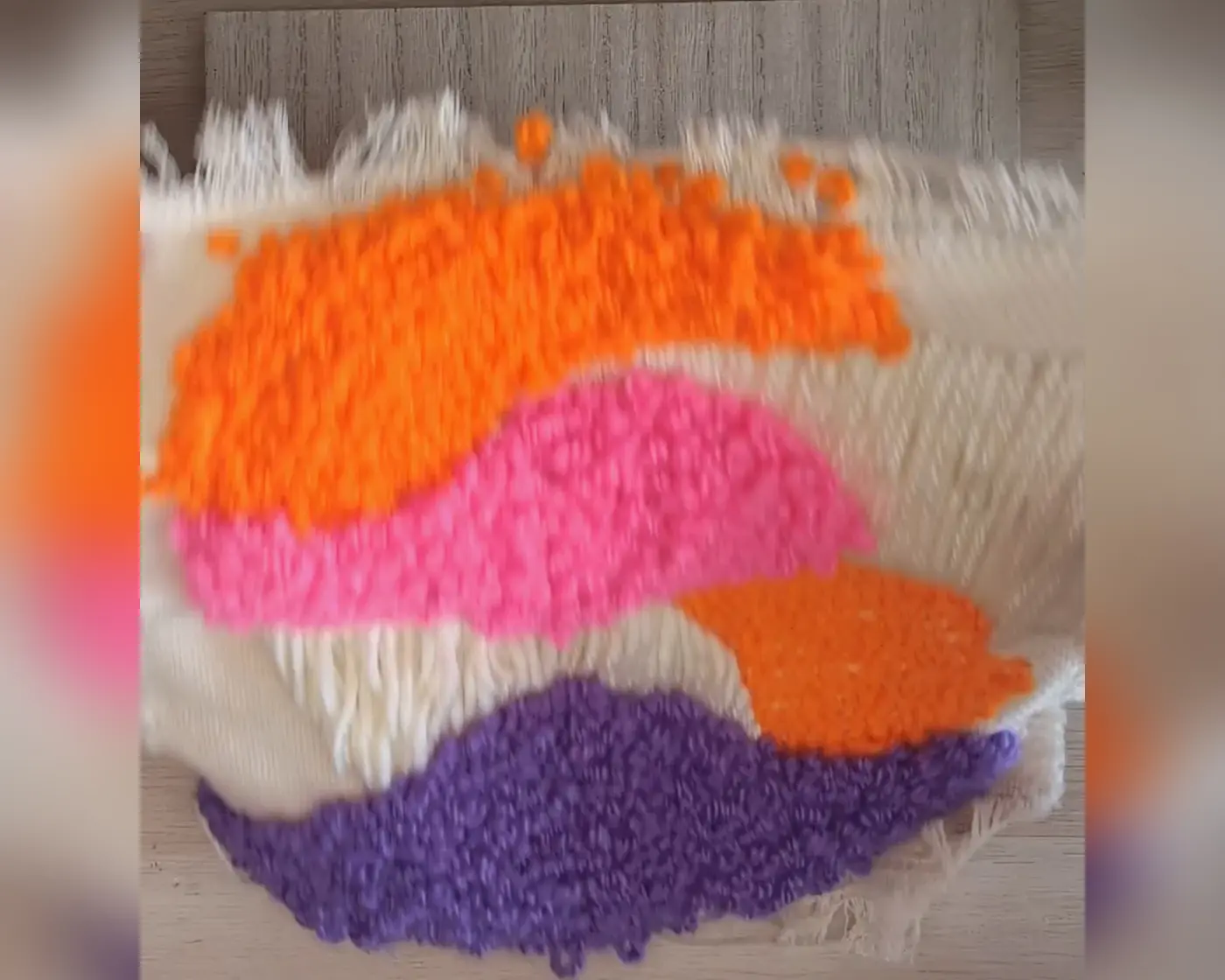 Voorkant van het punch needle werkstuk buiten de ring met golvende vlakken in oranje, roze en paars garen op beige stof