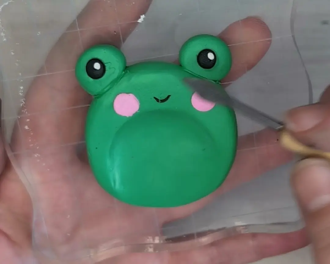 Handen gebruiken een fijne metalen spatel om een dun zwart kleimondje op het groene kikkergezichtje te boetseren, dat op een transparant ruitjesblok ligt.