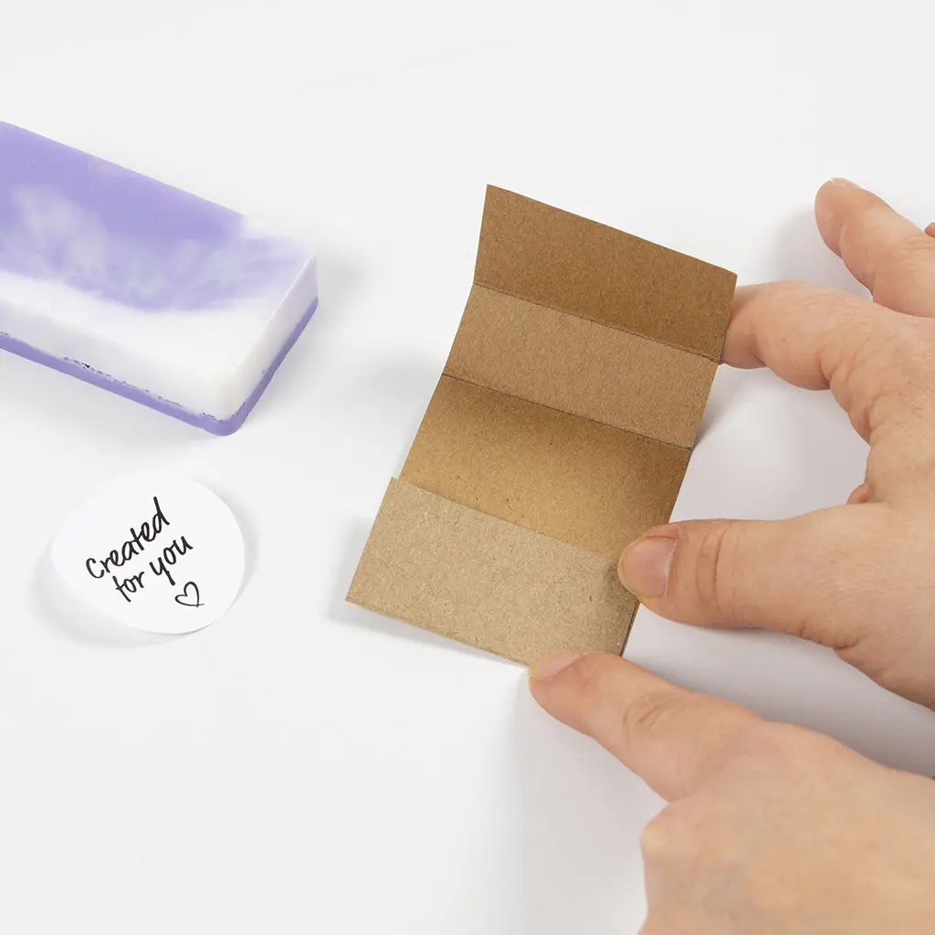 rechthoekig karton wordt gevouwen voor het label van de zeep, een ronde sticker ligt ernaast en de uitgeharde zeep erboven.