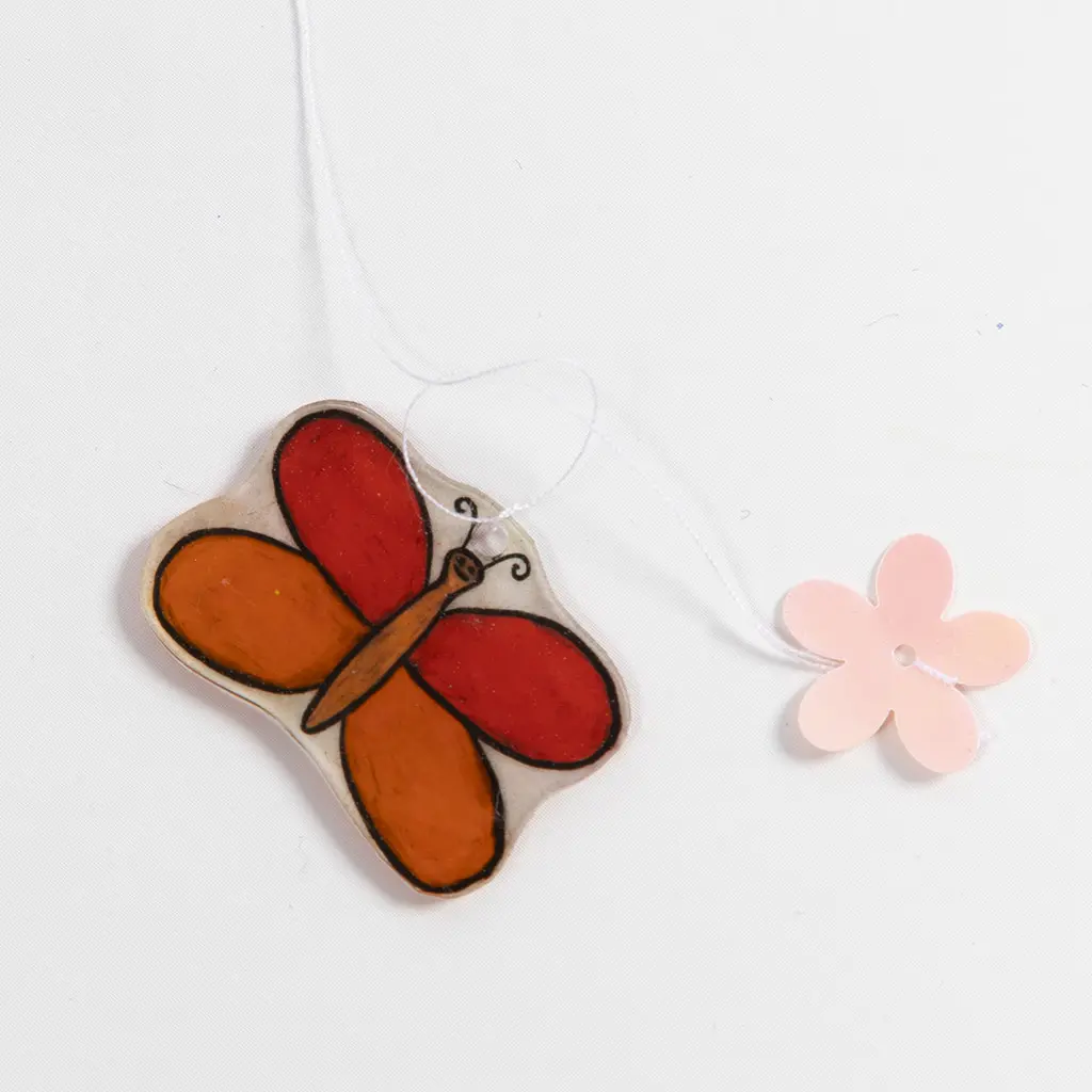 Een rood-oranje krimpfolievlinder en een lichtroze plastic bloemetje zijn aan een dunne witte draad geregen.
