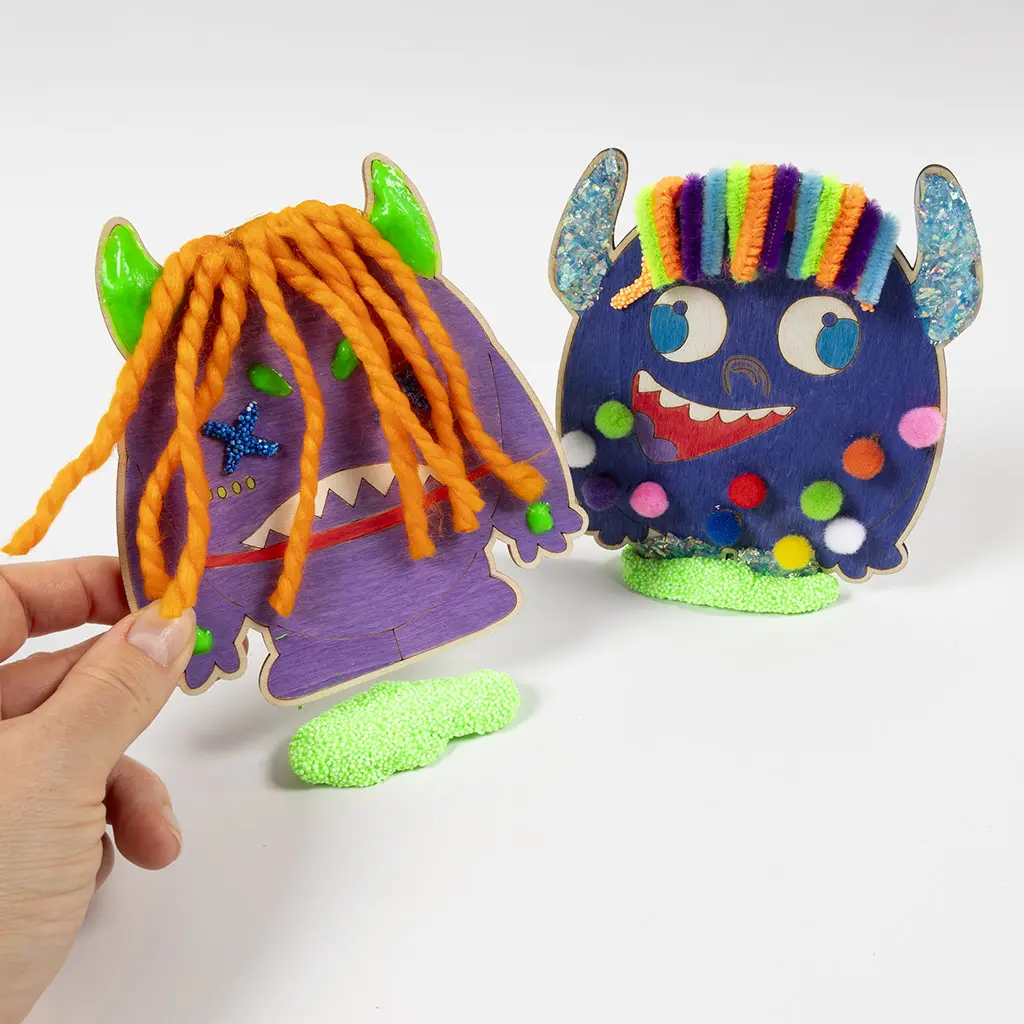 Een hand houdt het paarse houten monster rechtop boven een klompje felgroene Foam Clay. Op de achtergrond staat het blauwe monster al stevig rechtop in zijn eigen voetstuk van groene klei.