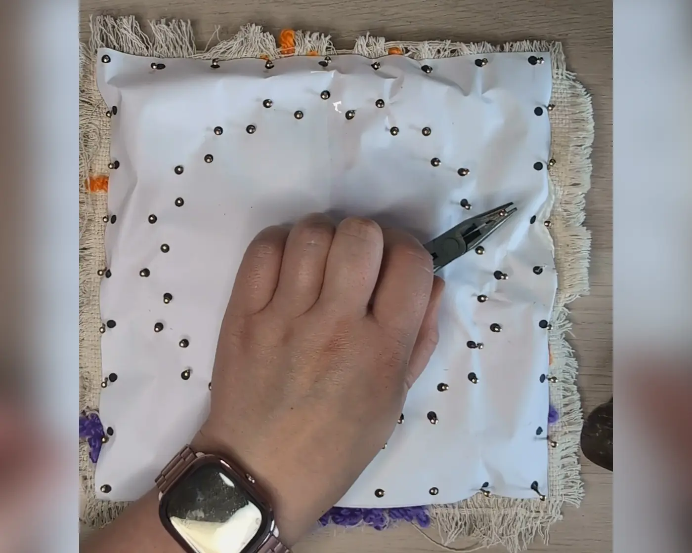 Hand die met een tang spijkers door het patroon in de punch needle stof slaat, met het patroon op de stof vastgezet
