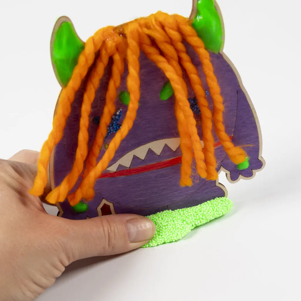 Close-up van een hand die de onderkant van het paarse houten monster in een langwerpig voetstuk van felgroene Foam Clay drukt, zodat het figuurtje zelfstandig kan blijven staan.