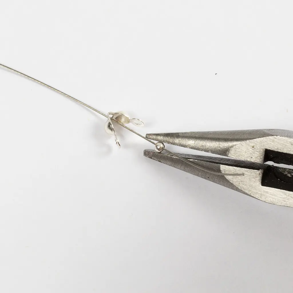 Een tang knijpt een klein zilveren knijpkraaltje dicht op een metaaldraad, vlak achter een openstaand zilveren kalotje (verkoopoogje).