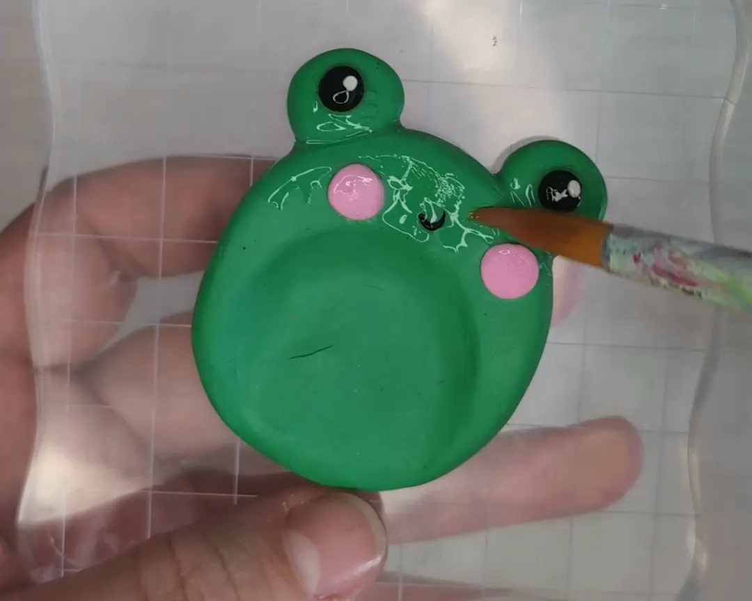 Een fijne kwast brengt een vloeibare, glanzende laklaag aan op de afgebakken groene kikker worry stone, waardoor de klei een beschermende glans krijgt.